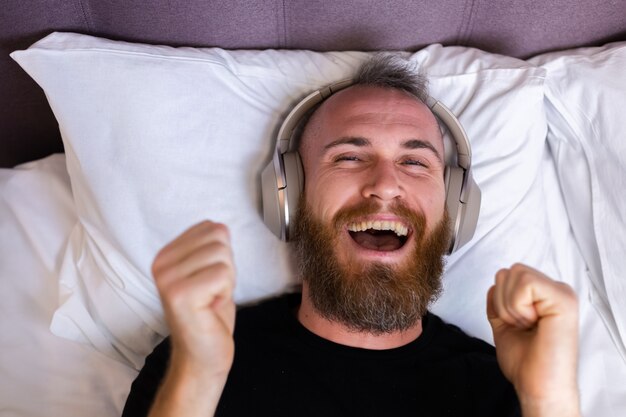 Feliz homem caucasiano na cama usando fone de ouvido ouve curtir sua música favorita, descansando sozinho, dançando.