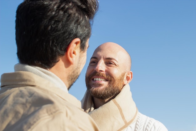 Feliz homem gay barbudo olhando namorado contra o céu azul. homem de meia idade sorridente no cachecol no encontro. conceito de homossexualidade