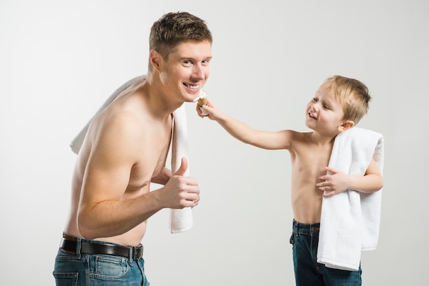 Feliz, homem jovem, mostrando, polegar cima, sinal, enquanto, seu, filho, aplicando, creme raspando, ligado, seu, rosto, contra, cinzento, fundo