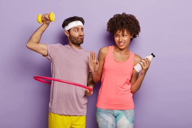 Feliz homem motivado com barba por fazer, gira bambolê, treina músculos com halteres e mulher afro insatisfeita faz gesto de recusa, segura garrafa de água