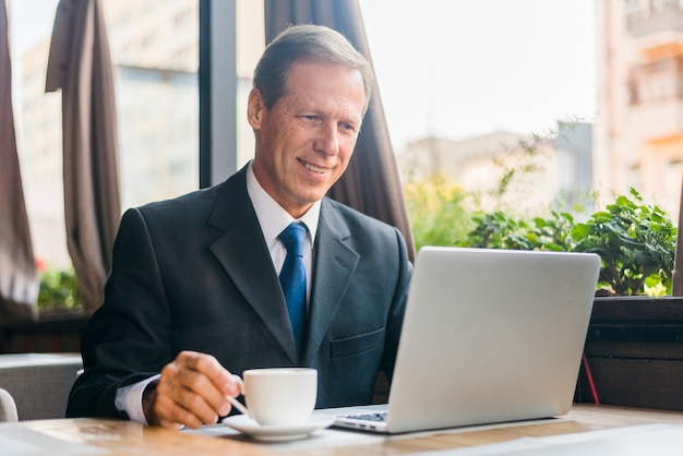 Feliz, homem negócios, usando computador portátil, com, xícara café, ligado, escrivaninha madeira