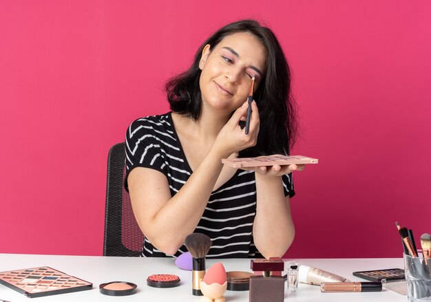 Feliz inclinando a cabeça jovem linda se senta à mesa com ferramentas de maquiagem, aplicação de sombra com pincel de maquiagem - isolado na parede rosa