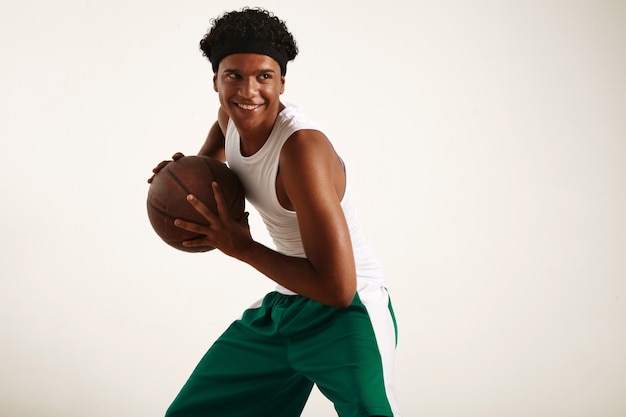 Feliz jogador de basquete preto com roupa verde e branco segurando uma bola de basquete marrom vintage, pose dinâmica em branco