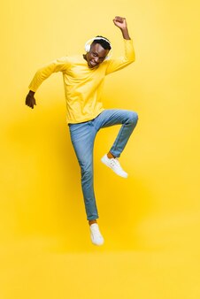 Feliz jovem africano usando fones de ouvido ouvindo música e pulando com a mão no fundo amarelo isolado do estúdio