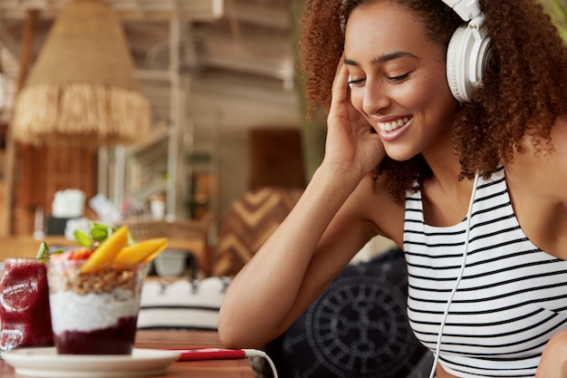 Foto grátis feliz jovem afro-americana com fones de ouvido pesquisa música no site da internet para fazer upload na lista de reprodução, usa um celular moderno, conectado a wi-fi em uma cafeteria aconchegante. menina hippie ouvindo áudio