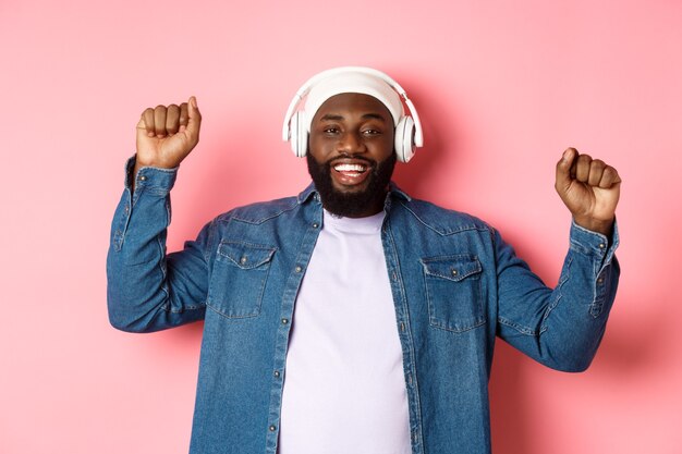 Feliz jovem afro-americano dançando e ouvindo música em fones de ouvido, erguendo os punhos e sorrindo, em pé sobre um fundo rosa