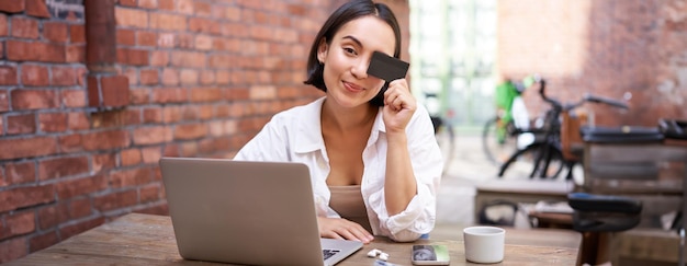 Foto grátis feliz jovem asiática sentada com laptop e mostrando cartão de crédito pagando contas on-line em compras
