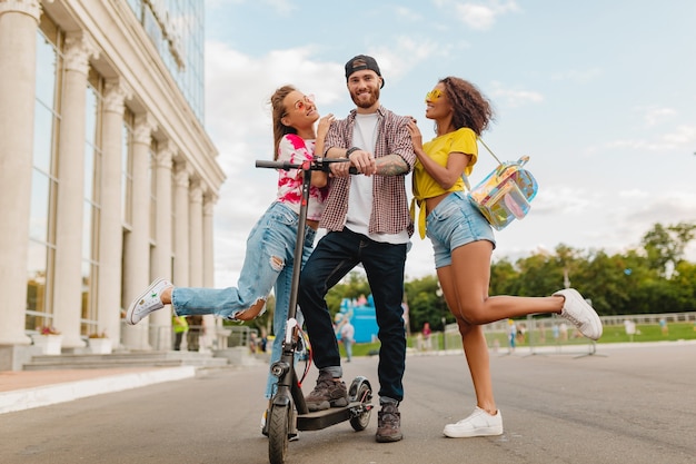 Feliz jovem companhia de amigos sorridentes andando na rua com patinete elétrica, homens e mulheres se divertindo juntos