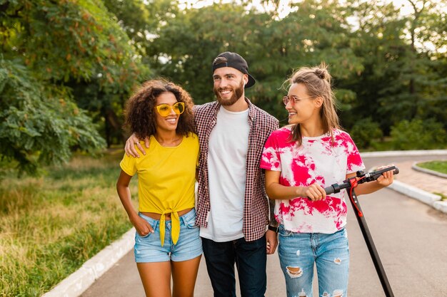 Feliz jovem companhia de amigos sorridentes caminhando no parque com patinete elétrica, homens e mulheres se divertindo juntos