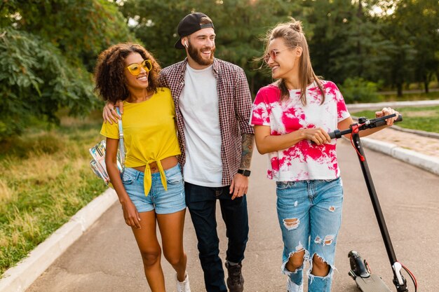 Feliz jovem companhia de amigos sorridentes caminhando no parque com patinete elétrica, homens e mulheres se divertindo juntos