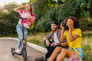 Foto grátis feliz jovem companhia de amigos sorridentes sentados no parque na grama com patinete elétrica, homens e mulheres se divertindo juntos