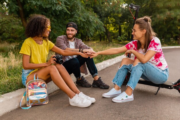 Feliz jovem companhia de amigos sorridentes sentados no parque na grama com patinete elétrica, homens e mulheres se divertindo juntos