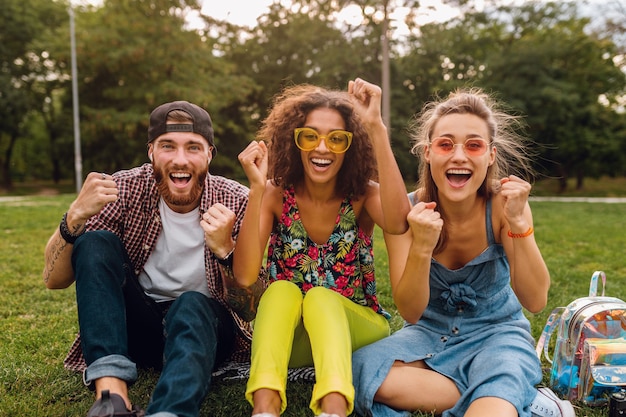 Feliz jovem companhia de amigos sorridentes sentados no parque na grama, homens e mulheres se divertindo juntos