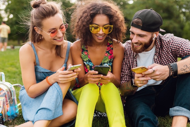 Feliz jovem companhia de amigos sorridentes sentados no parque usando smartphones, homens e mulheres se divertindo juntos