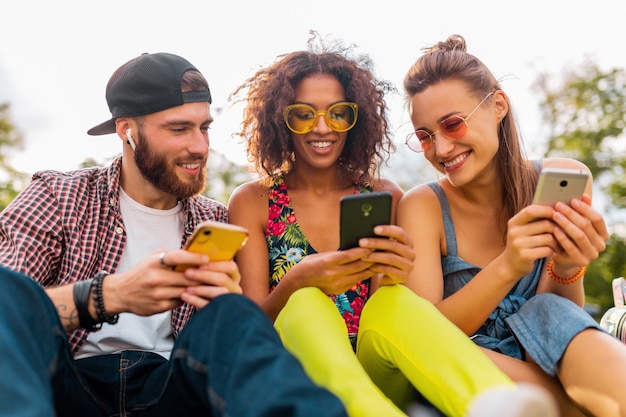 Feliz jovem companhia de amigos sorridentes sentados no parque usando smartphones, homens e mulheres se divertindo juntos