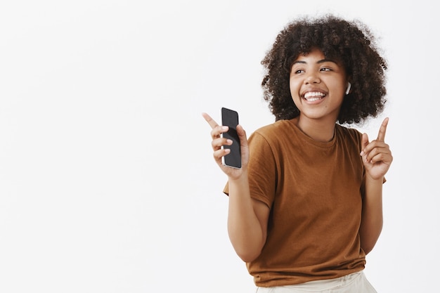 Feliz jovem dançarina afro-americana com uma camiseta marrom ouvindo música em fones de ouvido sem fio dançando e movendo os dedos no ritmo segurando um smartphone olhando para a esquerda com um sorriso