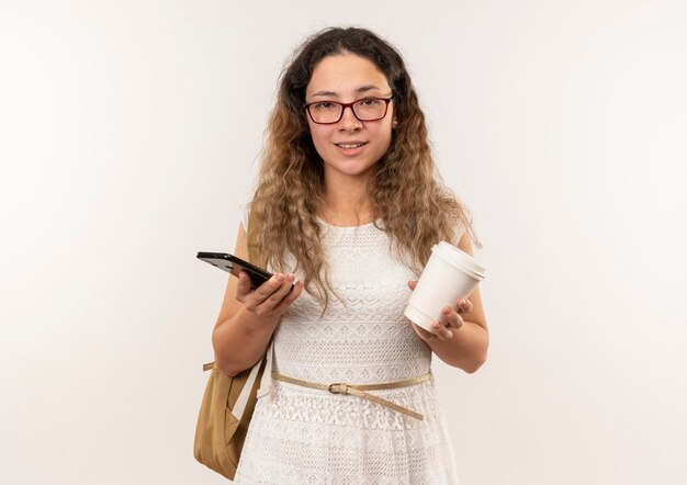 Feliz, jovem e bonita colegial de óculos e bolsa com as costas segurando a xícara de café e o celular isolado na parede branca