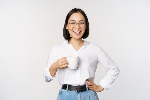 Feliz jovem e enérgica mulher asiática sorrindo bebendo segurando uma xícara de café em pé confiante contra o fundo branco