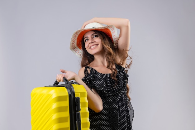 Feliz, jovem e linda garota viajante em um vestido de bolinhas com chapéu de verão, segurando a mala, olhando para cima sorrindo alegremente feliz e positiva em pé sobre um fundo branco
