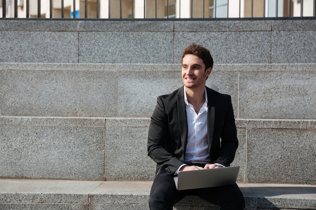 Feliz jovem empresário sentado ao ar livre usando laptop