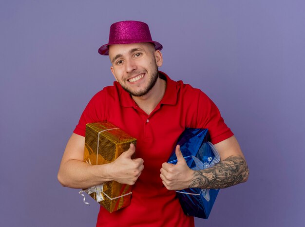 Feliz jovem eslavo bonito com chapéu de festa segurando embalagens de presente, olhando para a câmera isolada no fundo roxo