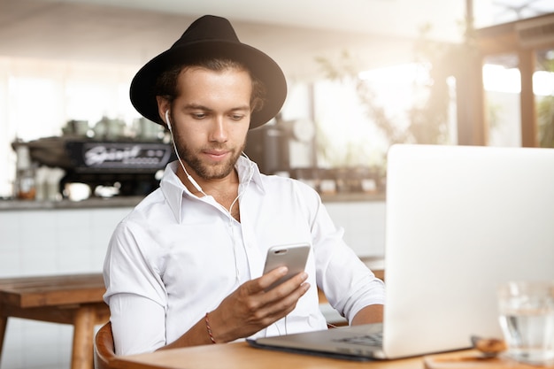 Feliz jovem estudante usando um chapéu estiloso e fones de ouvido, ouvindo música em seu celular, enquanto relaxa dentro de casa, na aconchegante cafeteria, sentado à mesa com o computador laptop.