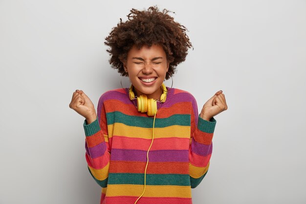 Feliz jovem garota afro cerra os punhos, comemora o momento agradável, ri agradavelmente, sorri amplamente, usa fones de ouvido no pescoço