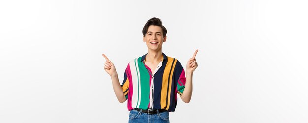 Feliz jovem gay apontando os dedos para o lado mostrando duas opções e sorrindo em pé contra o branco