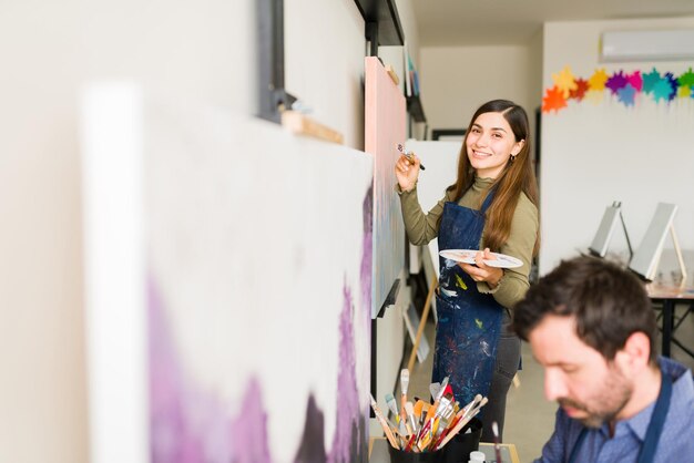 Feliz jovem hispânica com um avental está sorrindo enquanto pinta em uma tela grande para sua aula de arte para adultos