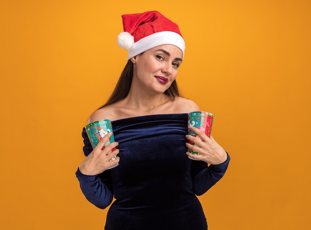 Feliz jovem linda com vestido azul e chapéu de Natal segurando copos de Natal isolados em fundo laranja