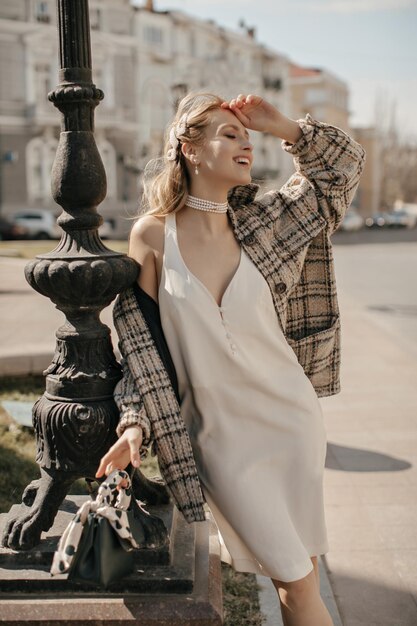 Feliz jovem loira de vestido branco moderno e casaco xadrez de tweed sorri sinceramente e posa na praça da cidade