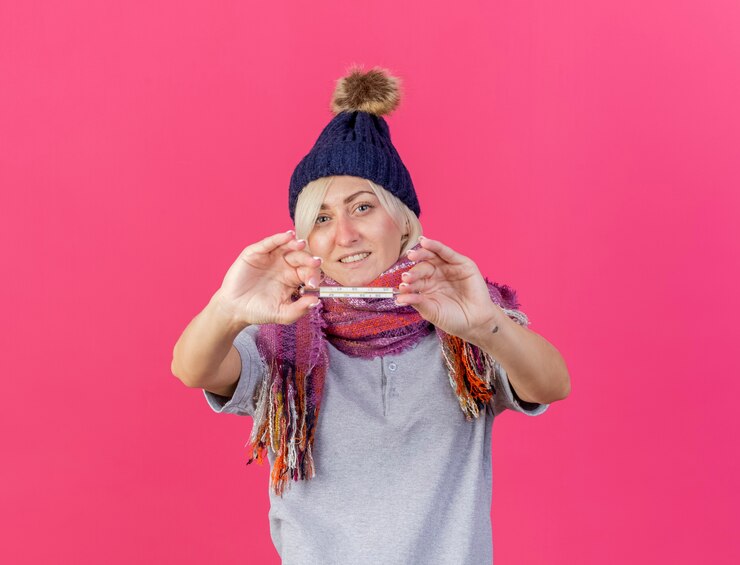Foto grátis feliz jovem loira doente eslava usando chapéu de inverno e lenço segurando termômetro isolado na parede rosa com espaço de cópia