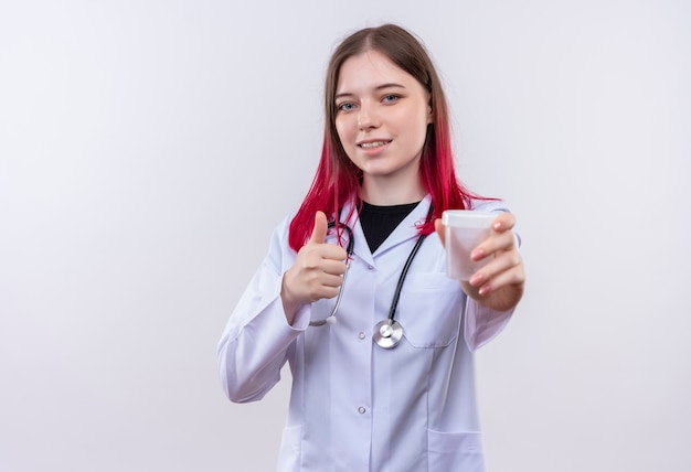 Feliz jovem médica usando estetoscópio manto médico, segurando a lata vazia com o polegar para cima, sobre fundo branco isolado