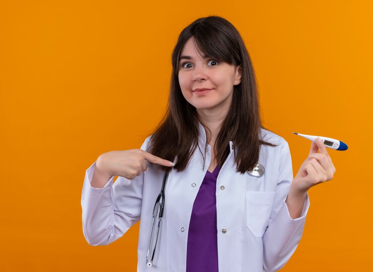 Foto grátis feliz jovem médica vestida de médico com estetoscópio segura o termômetro e aponta para o termômetro em um fundo laranja isolado com espaço de cópia