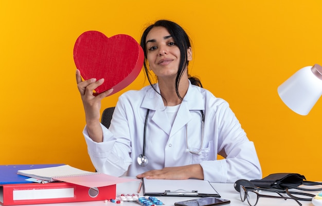 Foto grátis feliz jovem médica vestindo túnica médica com estetoscópio e sentada à mesa com ferramentas médicas segurando uma caixa em forma de coração isolada na parede amarela