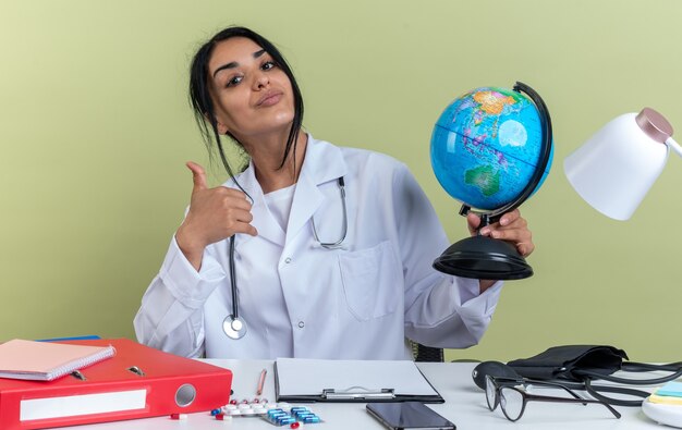 Feliz jovem médica vestindo túnica médica com estetoscópio sentada na mesa com ferramentas médicas segurando um globo aparecendo o polegar isolado na parede verde oliva