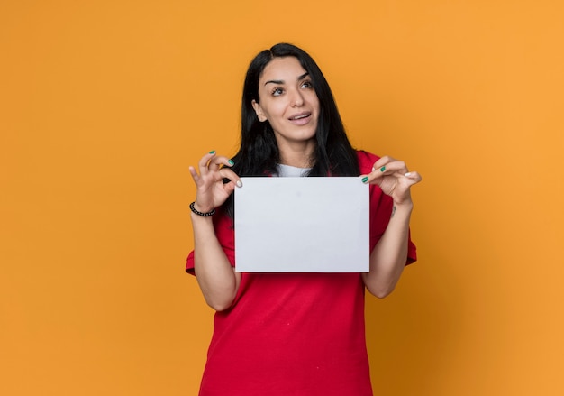 Foto grátis feliz, jovem morena caucasiana, vestindo uma camisa vermelha, segurando uma folha de papel em branco, olhando para o lado isolado na parede laranja