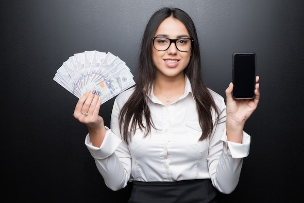 Feliz jovem morena de camisa branca mostrando smartphone com tela em branco e dinheiro nas mãos isoladas na parede preta