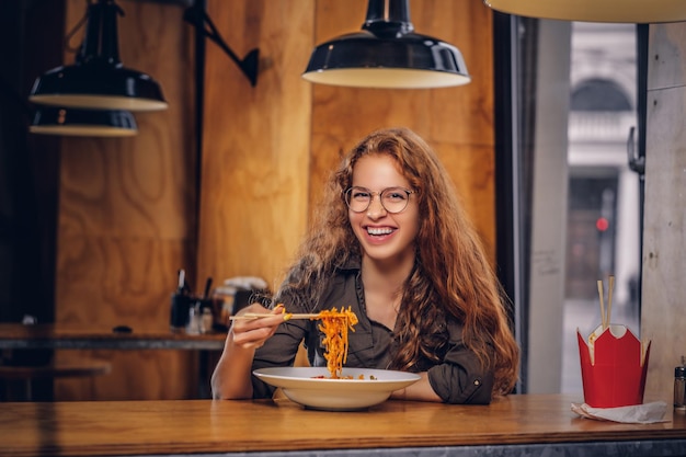 Feliz jovem ruiva vestindo roupas casuais e óculos comendo macarrão picante em um restaurante asiático.