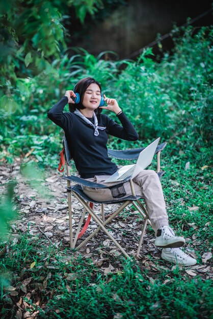 Feliz jovem sentada na cadeira de acampamento usando fone de ouvido ouvindo música do computador portátil enquanto relaxa no acampamento na floresta