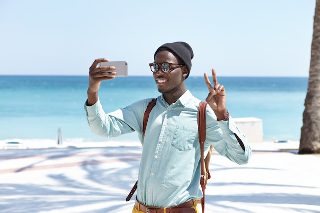 Feliz jovem viajante com mochila olhando e sorrindo para a câmera em seu smartphone