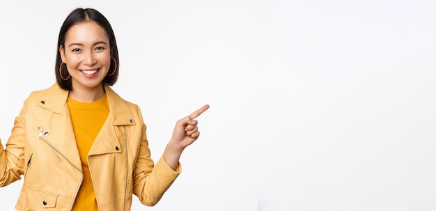 Foto grátis feliz linda garota asiática apontando os dedos para a esquerda e para a direita mostrando o logotipo do banner demonstrando a venda em pé na jaqueta amarela sobre fundo branco