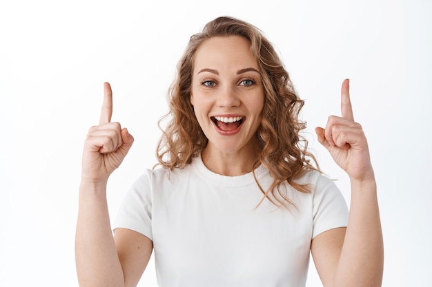 Feliz linda garota com cabelo loiro encaracolado, apontando os dedos para cima e sorrindo satisfeita, mostrando um anúncio, em pé sobre uma parede branca