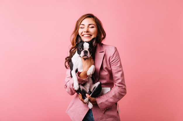 Feliz linda garota segurando bulldog francês e sorrindo com os olhos fechados. Retrato de mulher ruiva romântica com cachorrinho isolado em rosa.
