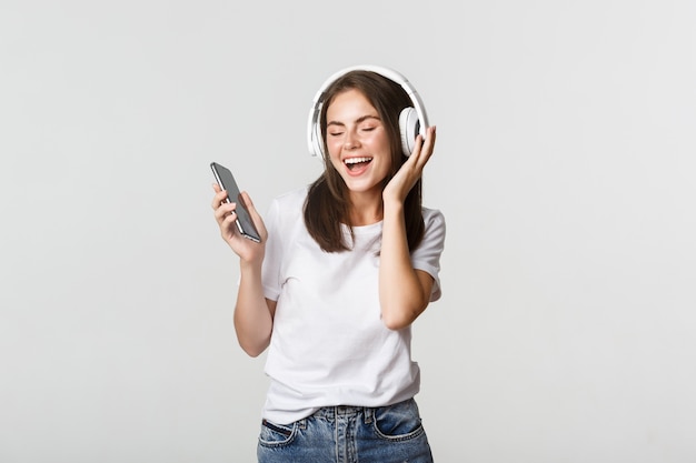 Feliz linda menina morena dançando e ouvindo música em fones de ouvido sem fio, segurando o smartphone.