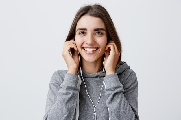 Feliz linda morena jovem caucasiana cabelos longos com capuz cinza, sorrindo com dentes, segurando os fones de ouvido com as mãos, com expressão de rosto animado e alegre