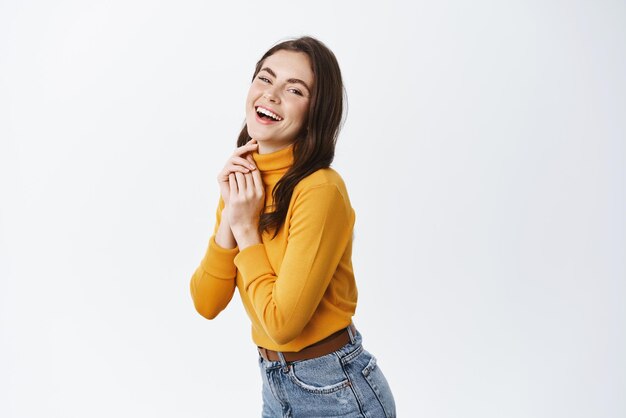 Feliz linda mulher de suéter amarelo rindo e expressando emoções positivas sorrindo para a câmera em pé meio virada no fundo branco