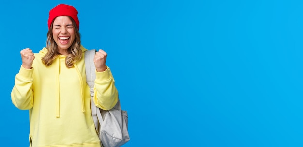 Foto grátis feliz loira alegre de gorro vermelho e capuz amarelo fecha os olhos aliviados e sorrindo triunfando