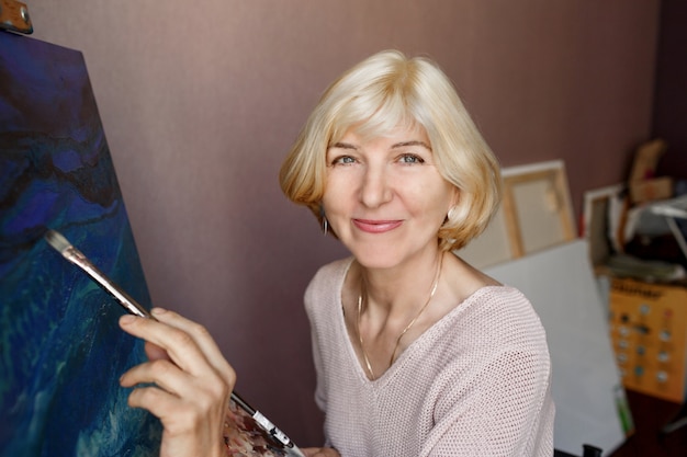 Foto grátis feliz loira madura pintura feminina em tela em casa