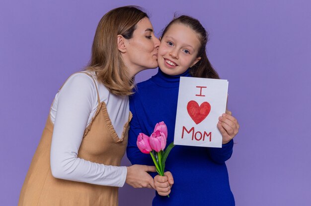 Feliz mãe beijando-a sorridente e feliz pastora segurando um cartão e flores de tulipas comemorando o dia internacional da mulher em pé sobre a parede roxa